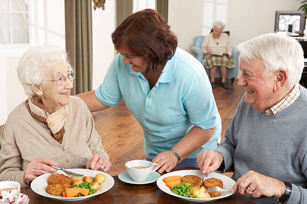 Senior Couple Being Served Meal By Carer In Care Home